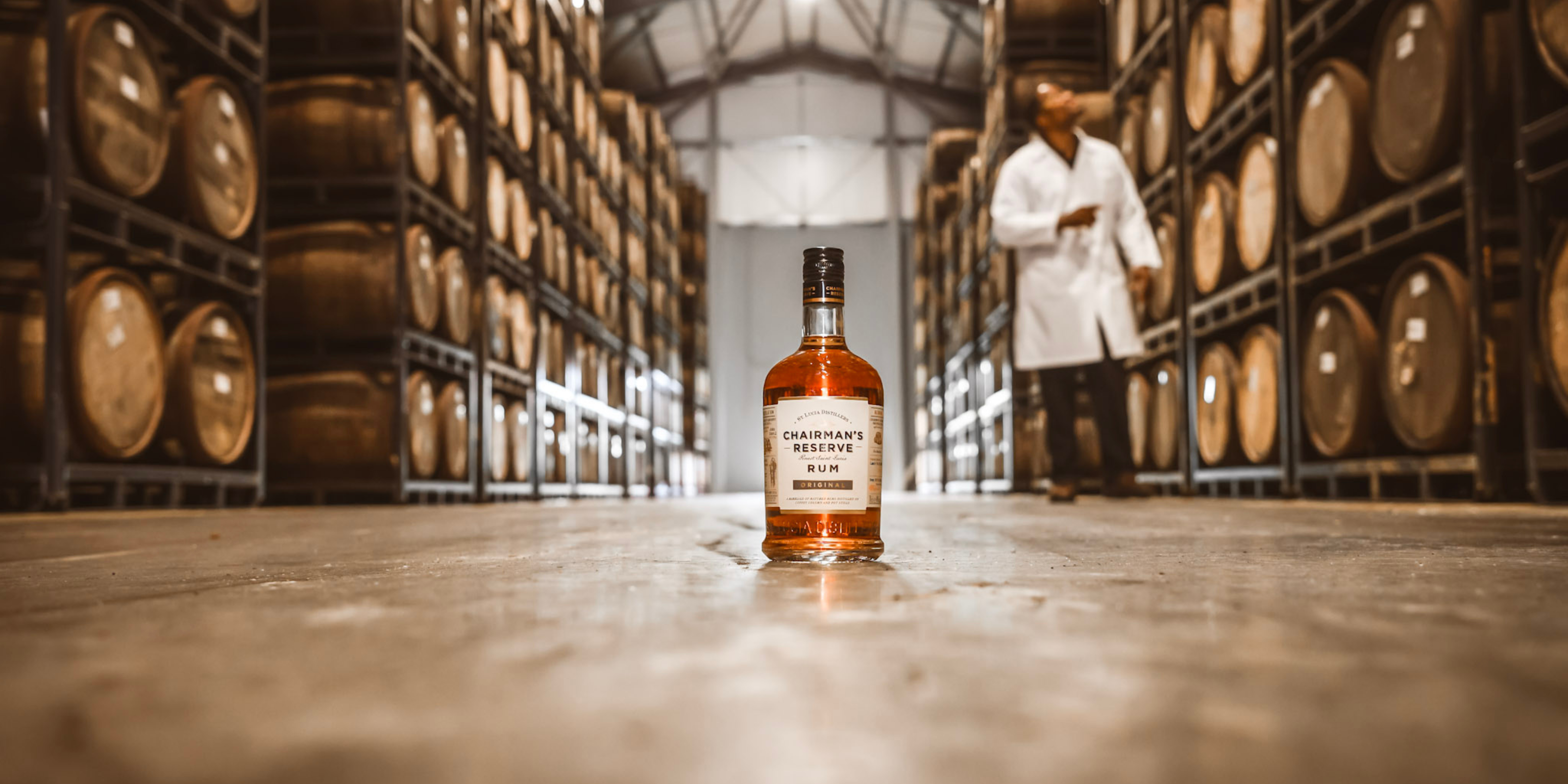 Bottle of Chairman’s Reserve Rum displayed in the distillery aging warehouse, surrounded by oak barrels.