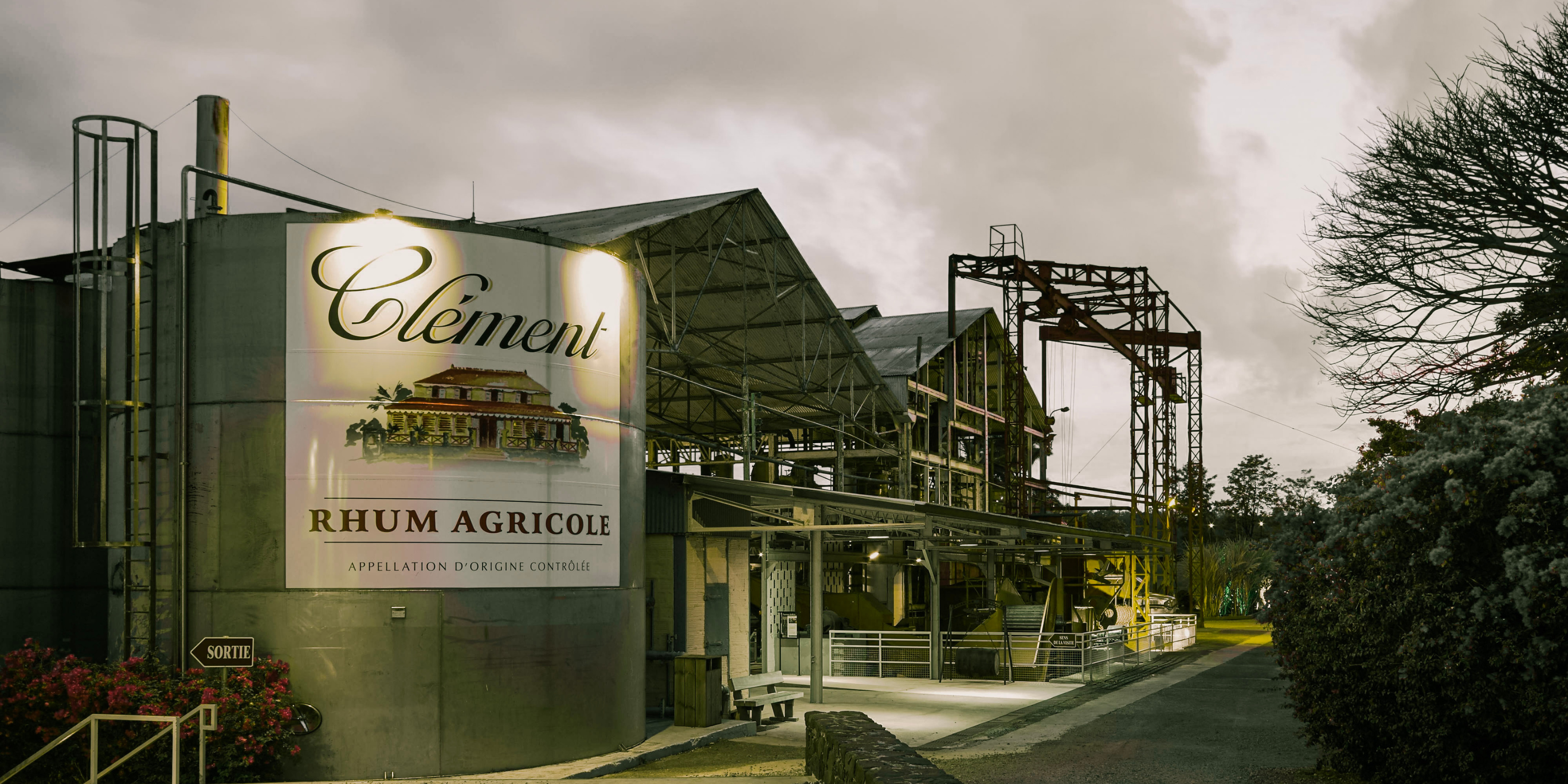 Exterior view of the Rhum Clément distillery in Martinique at dusk, featuring the illuminated Clément Rhum Agricole sign and the traditional rum production facility.