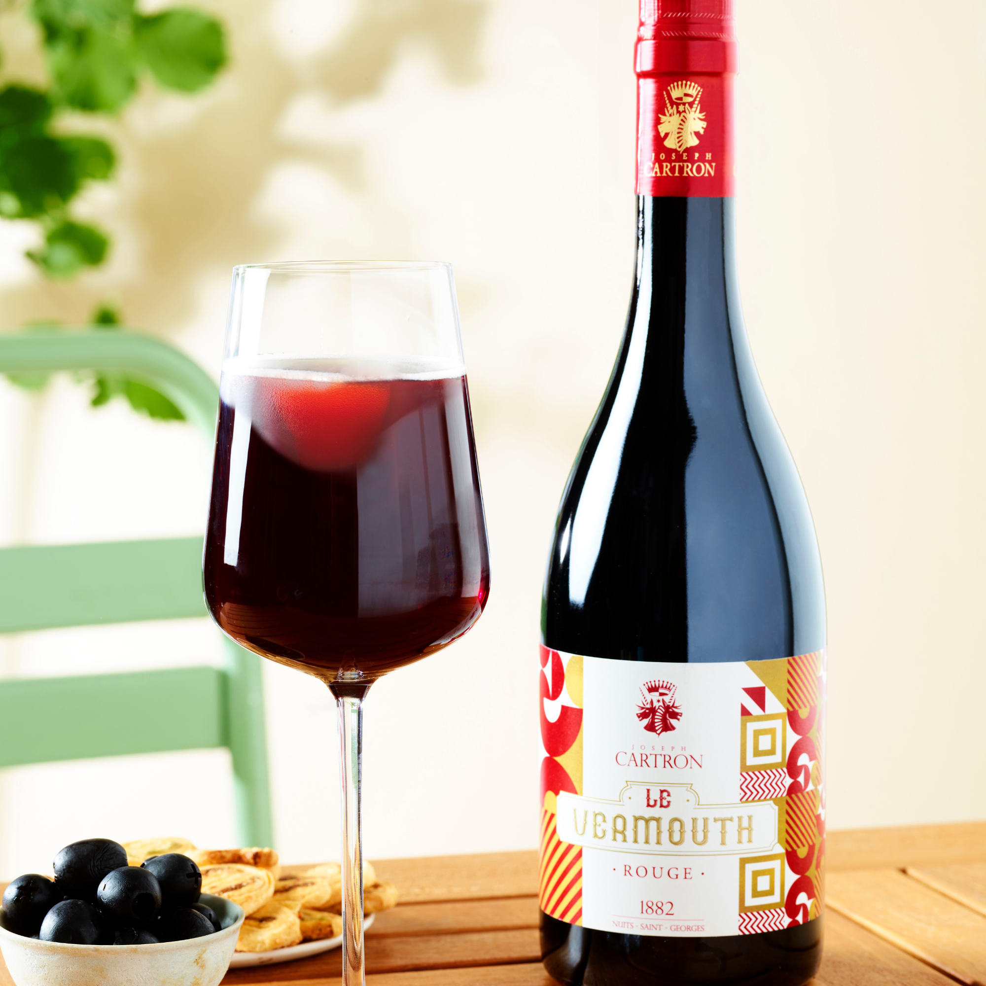 Bottle of Vermouth Rouge with a glass on a wooden table, surrounded by grapes and bread.