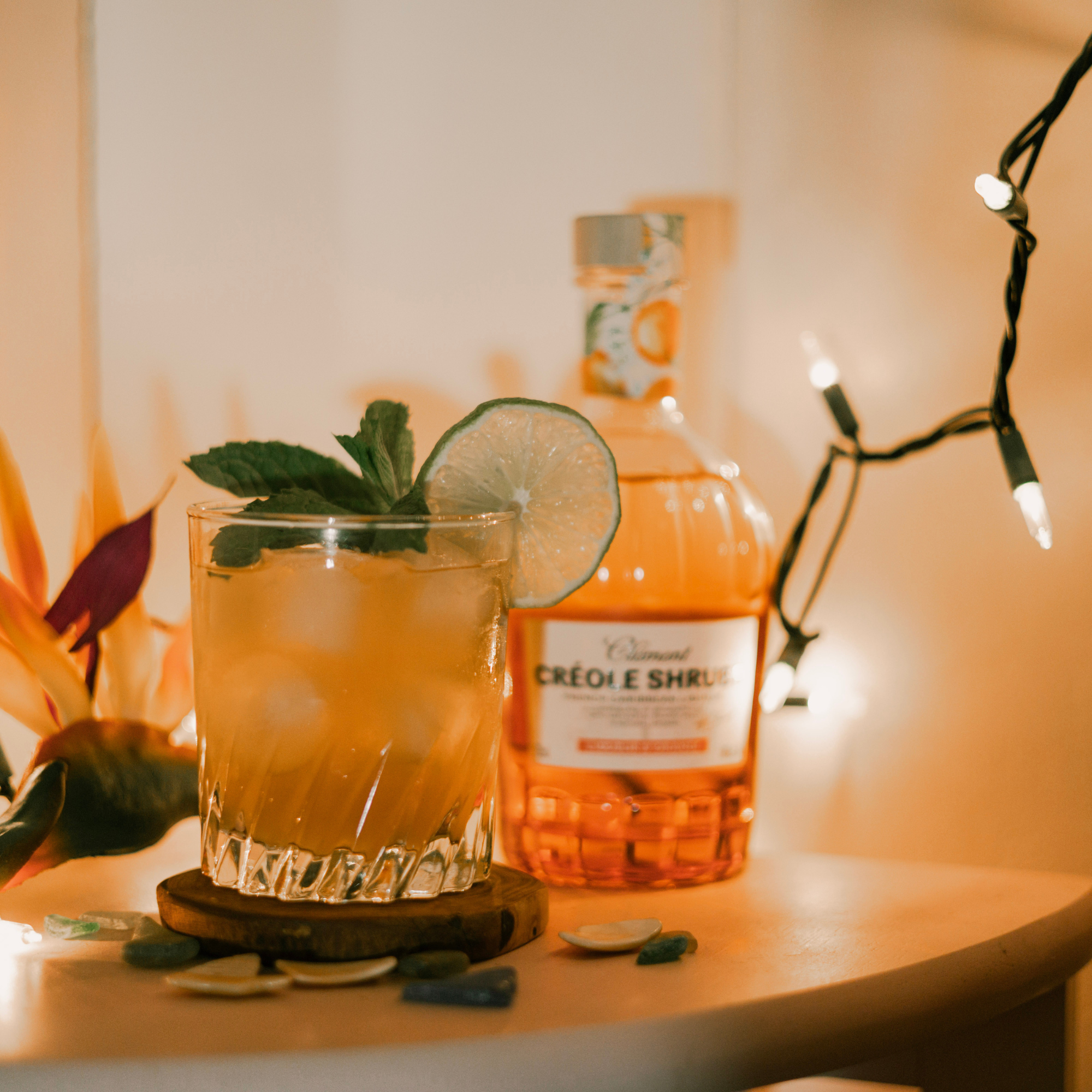 Glass of orange cocktail with lime and mint next to a bottle of Créole Shrubs on a table with string lights.