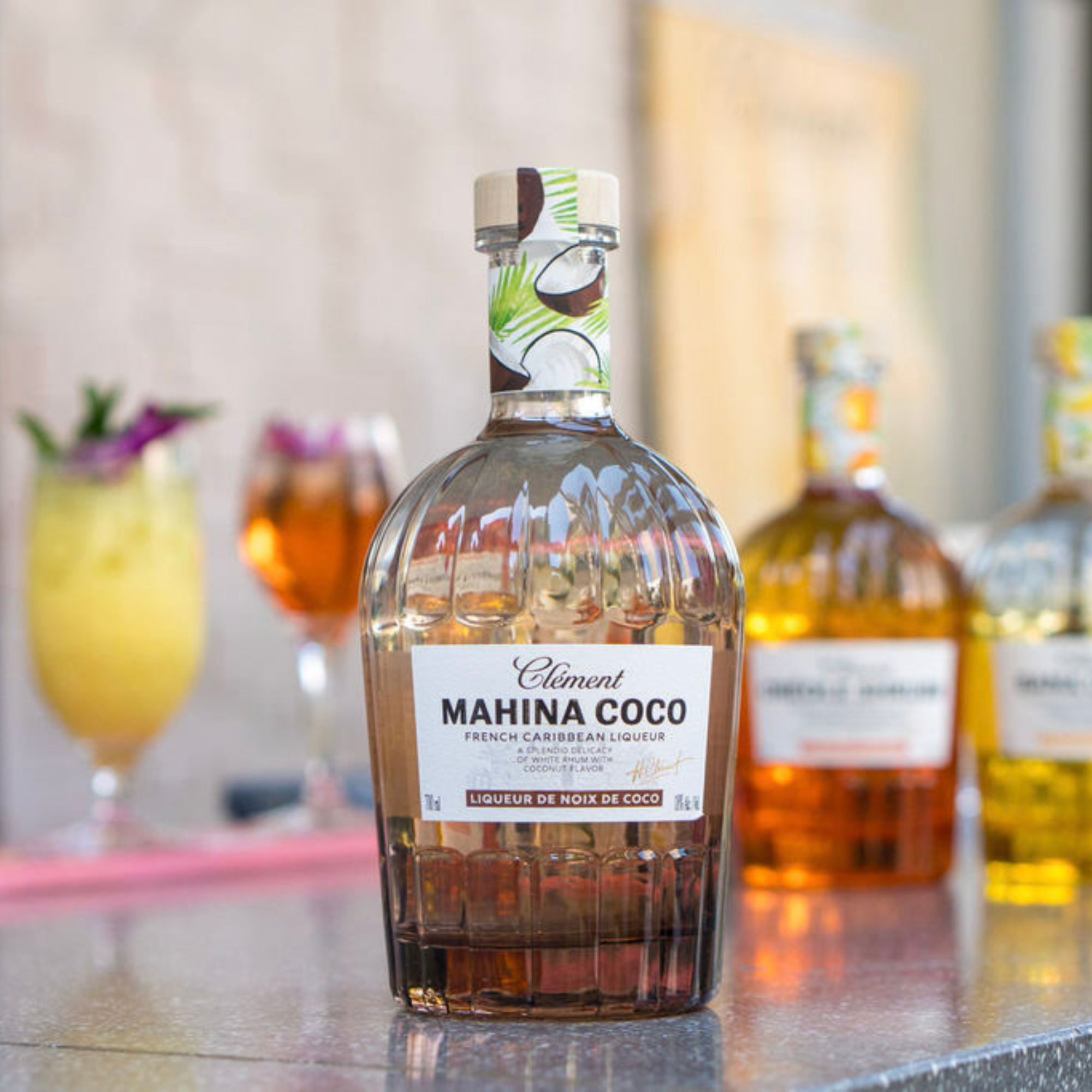 Bottle of Clément Mahina Coco liqueur on a bar counter with cocktails in the background.