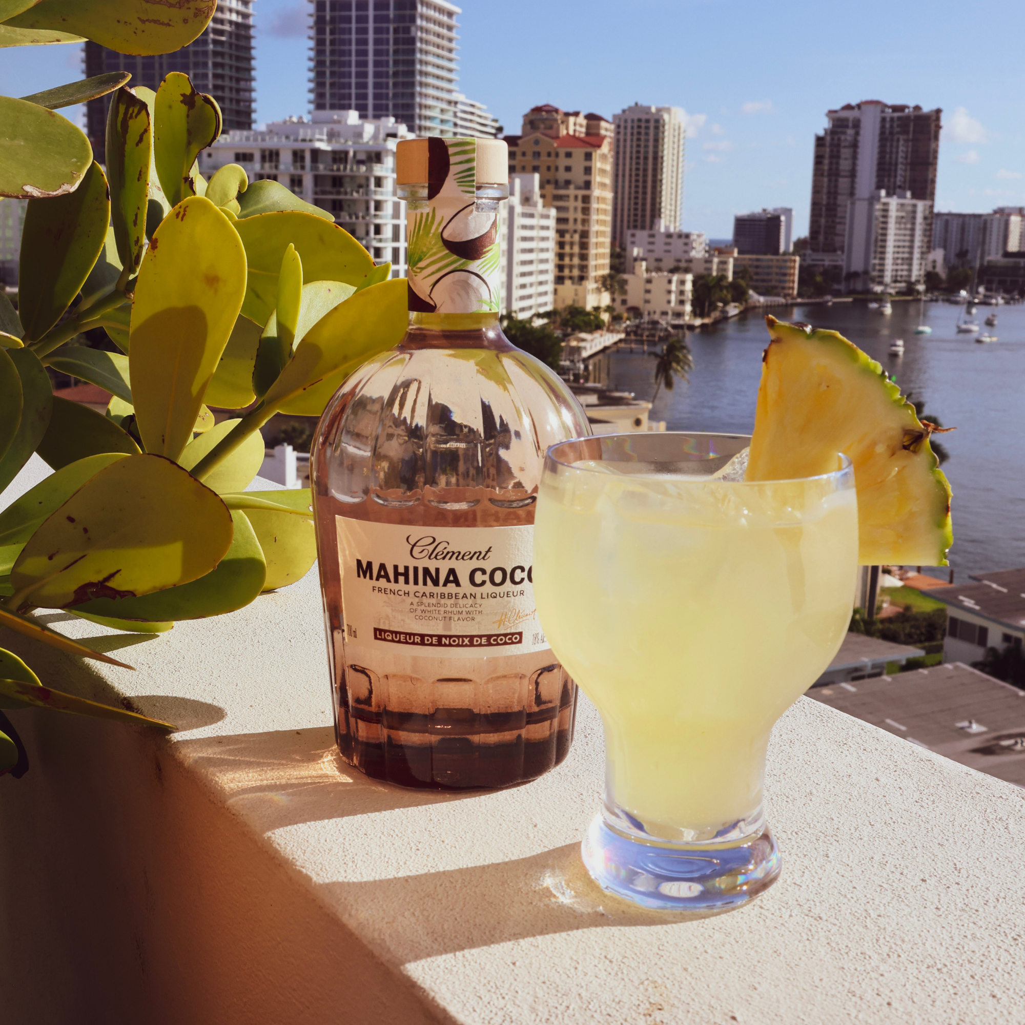 Bottle of Clément Mahina Coco with a cocktail garnished with a pineapple slice, set against a cityscape.