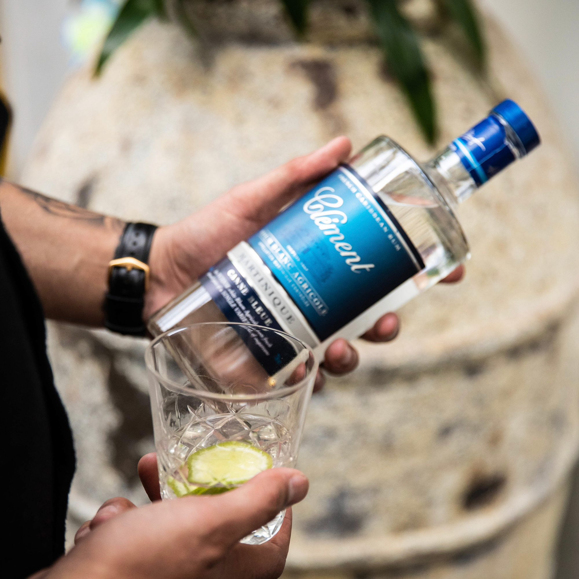 Person pouring a clear liquid from a bottle labeled 'Clement' into a glass with ice and a lime wedge.