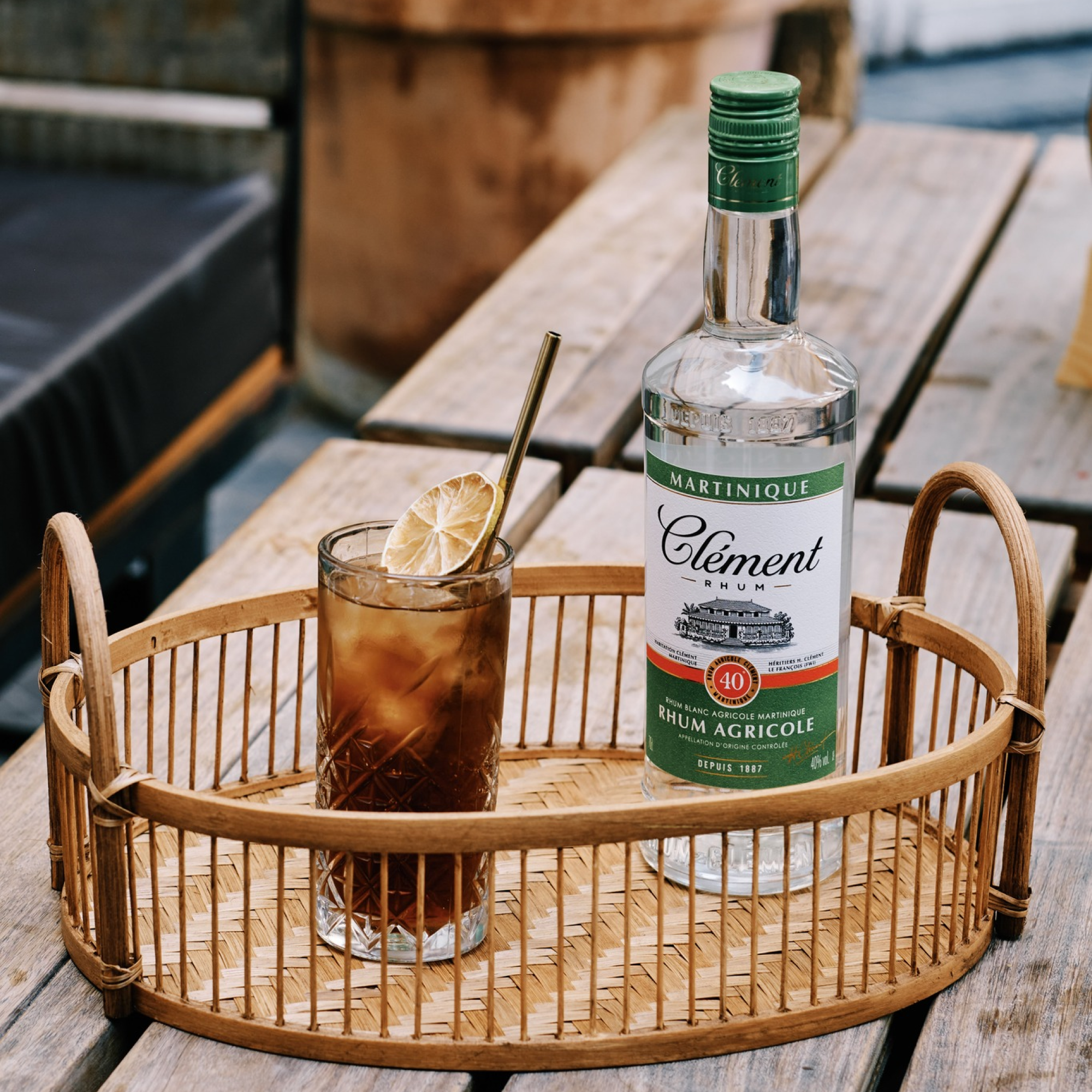Bottle of Clément Rhum with a cocktail on a wooden tray outdoors.