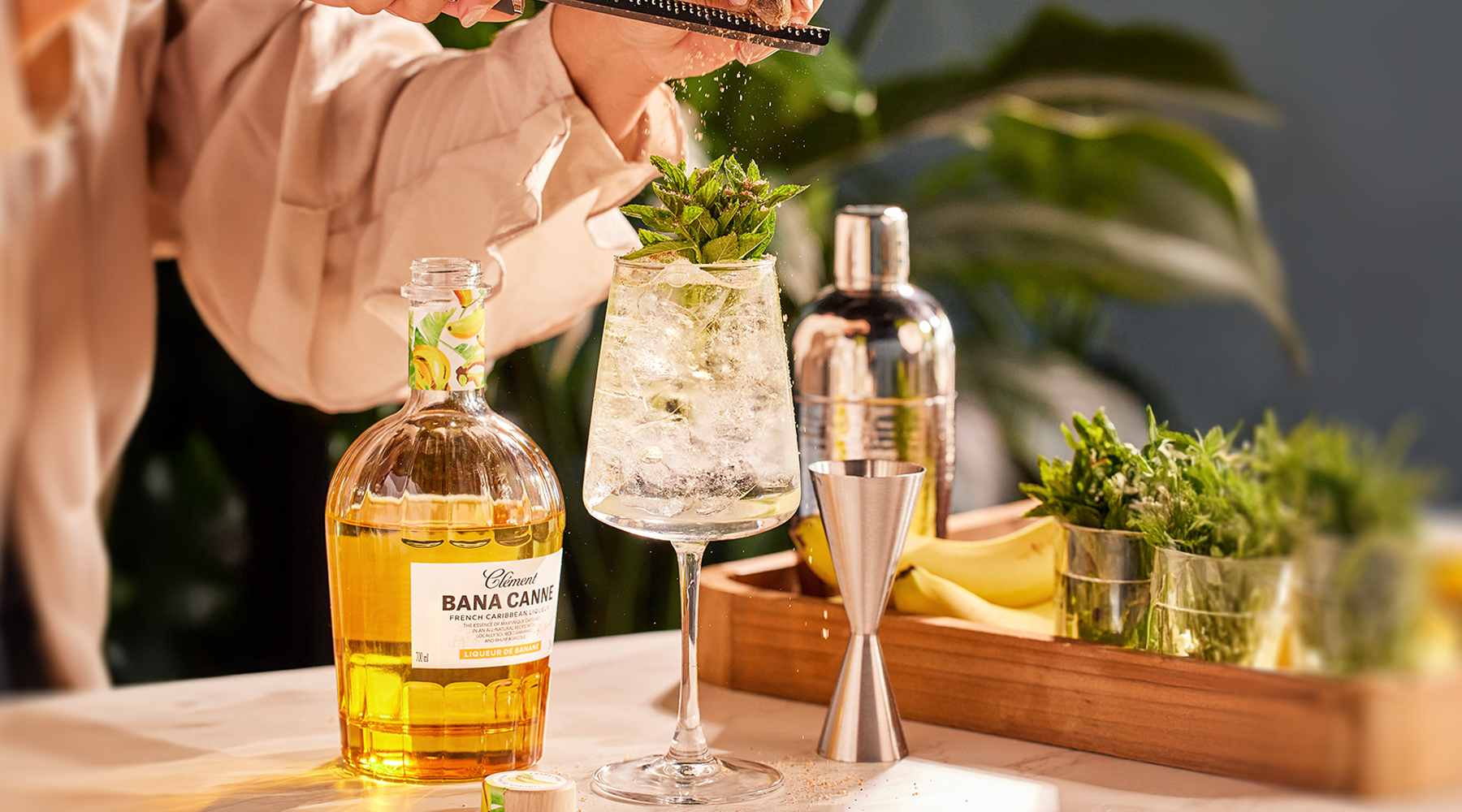 Person preparing a cocktail with Bana Canne liqueur and garnishes on a table.