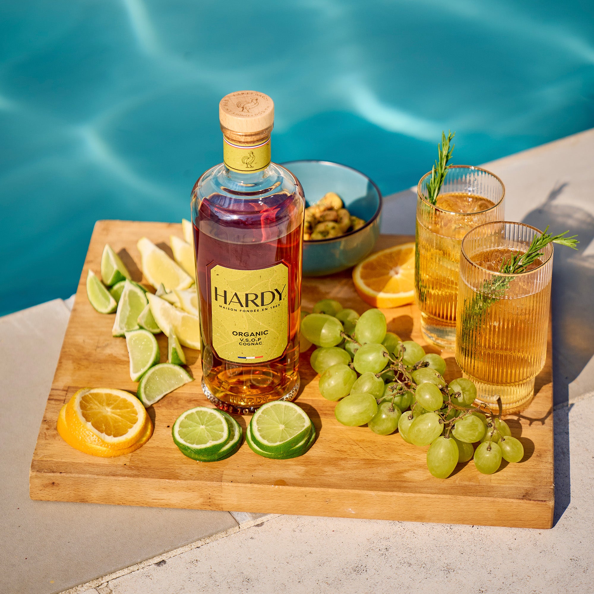 Hardy organic bottle on a wooden board with citrus fruits, grapes, and cocktails by a pool.