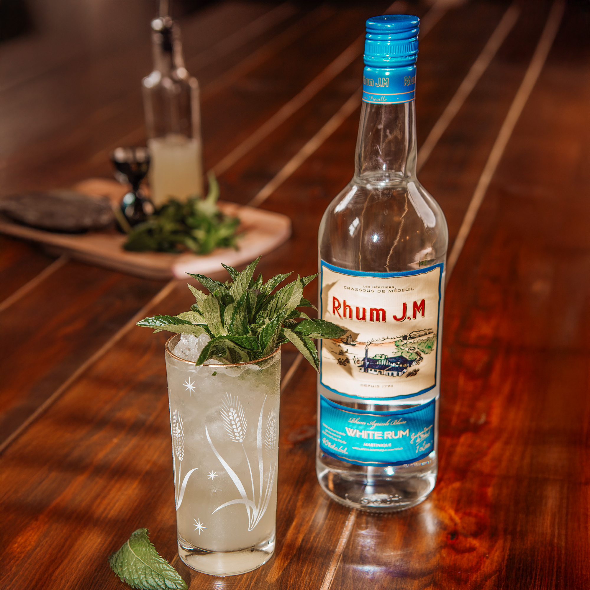Bottle of white Rhum J.M. rum on a wooden table with a cocktail garnished with mint leaves.