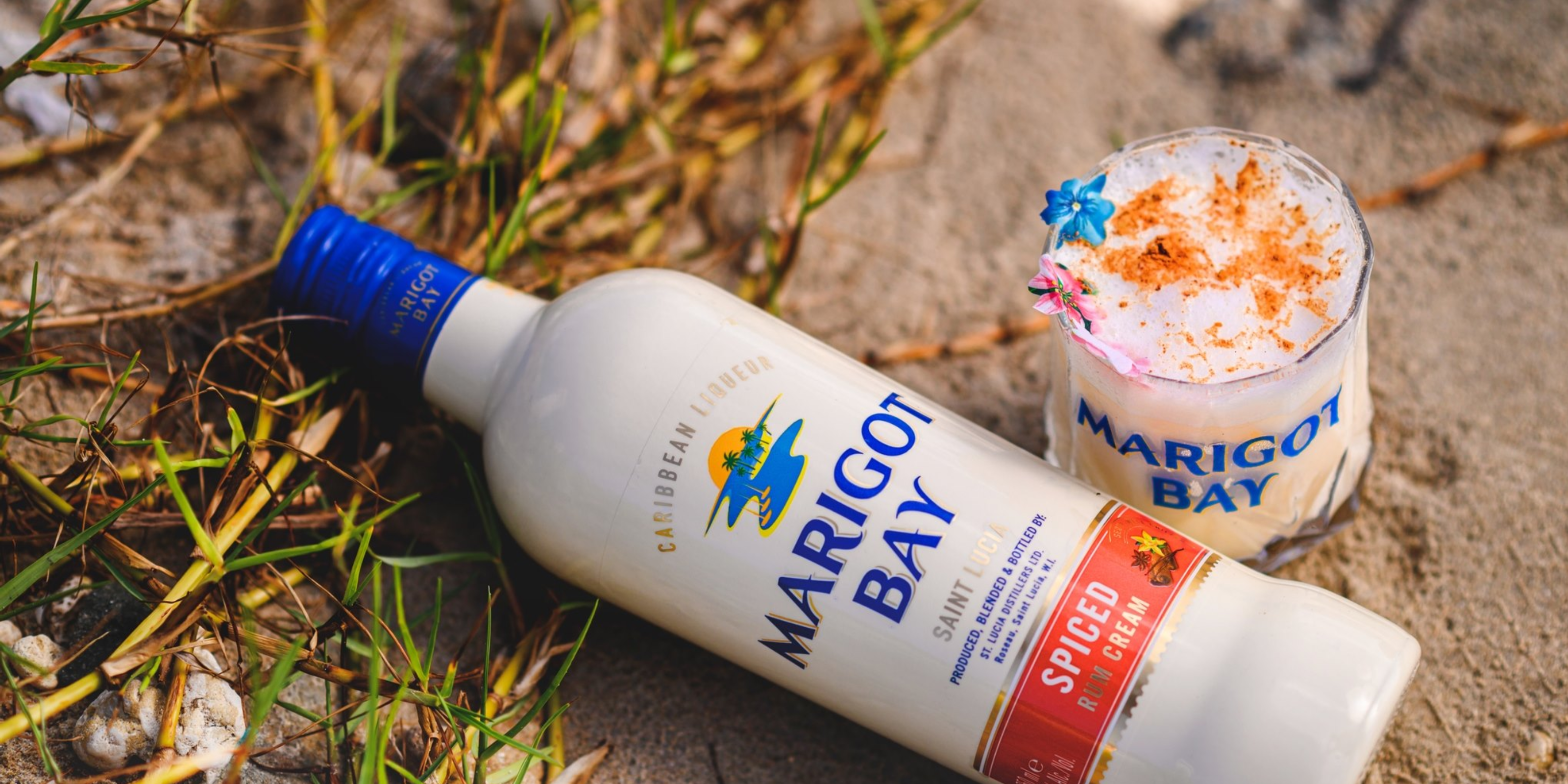 Bottle of Marigot Bay Spiced Rum Cream and a garnished cocktail resting on sandy beach grass in Saint Lucia.