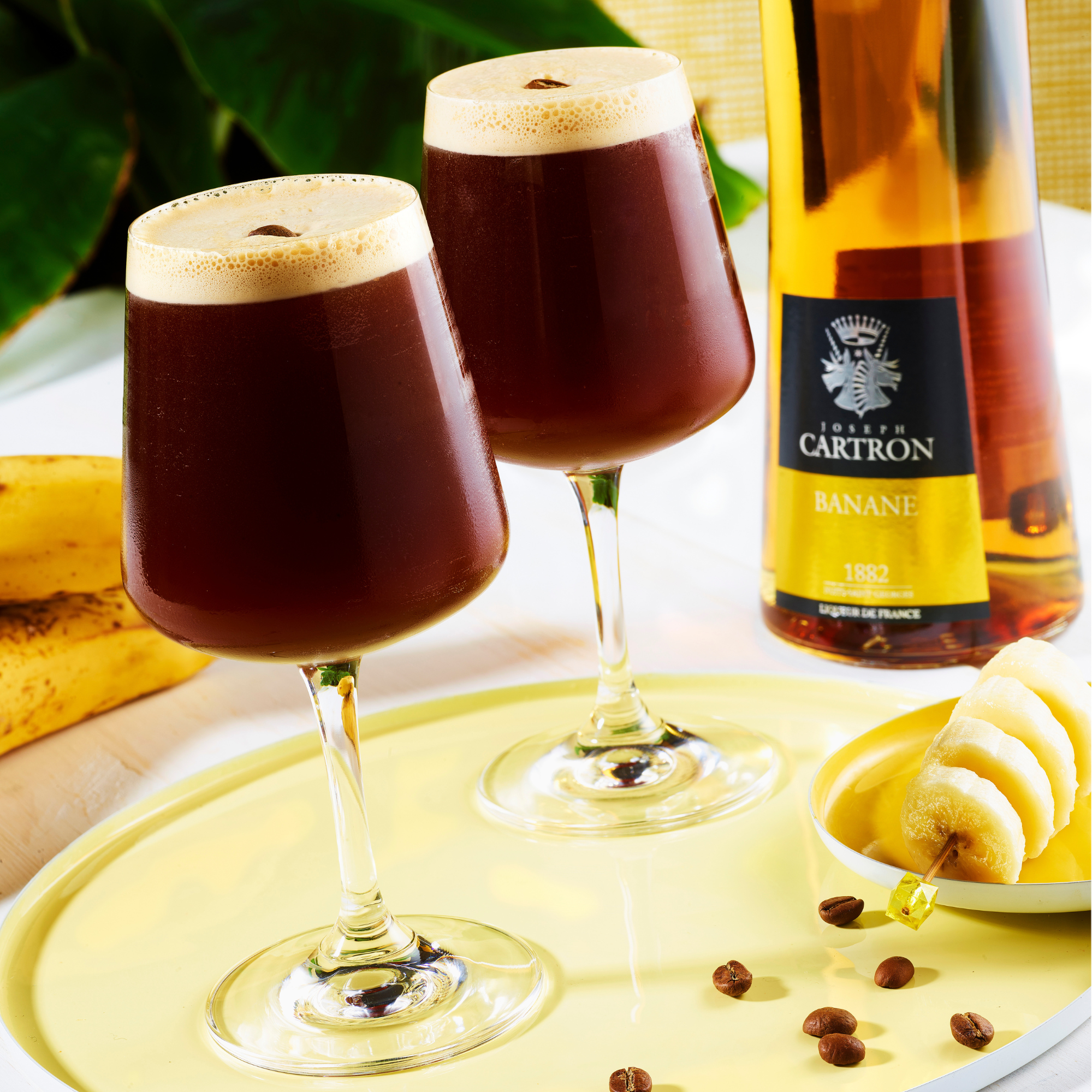 Two glasses of dark liquid with a bottle labeled 'Cartron Banane' on a yellow tray with bananas.