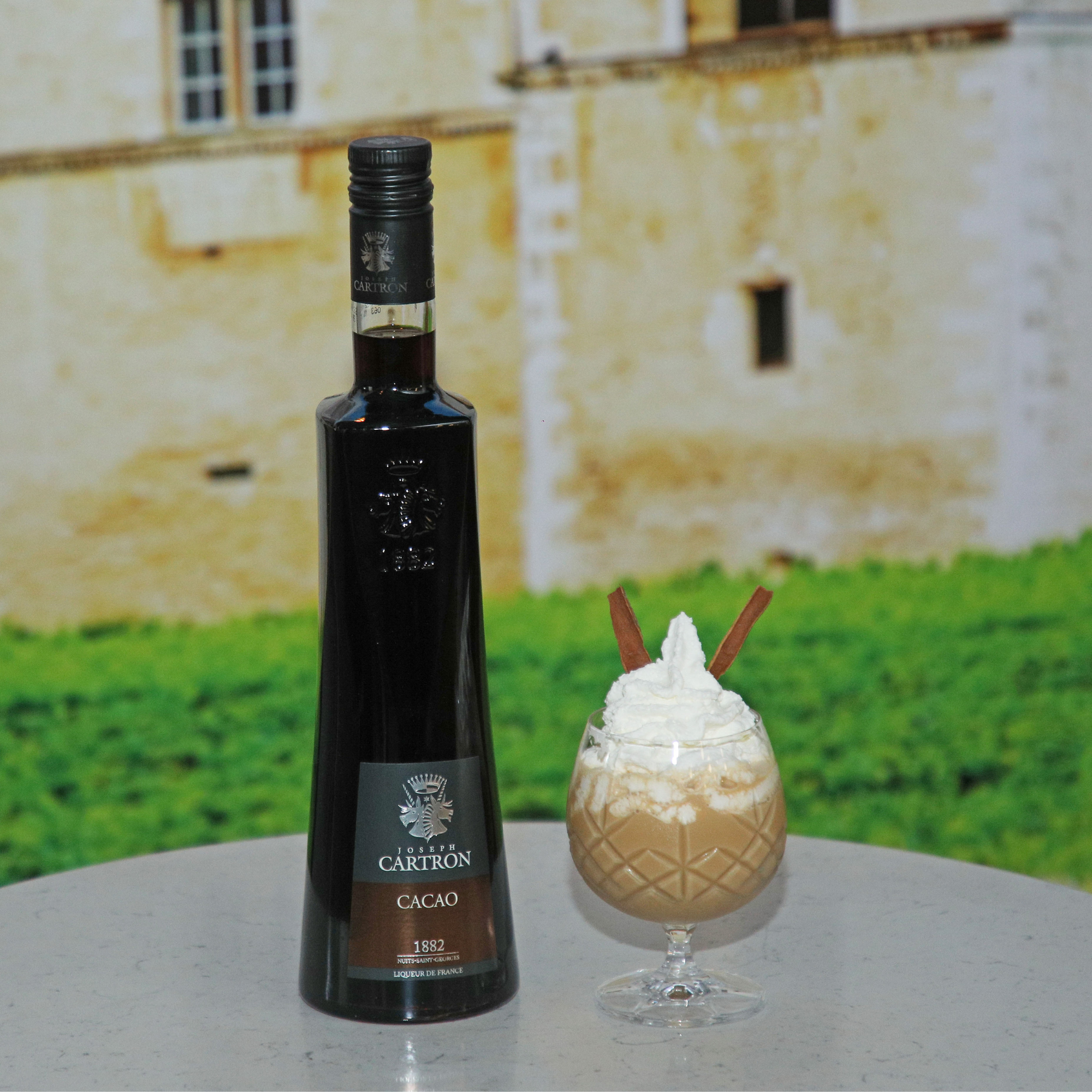 Bottle of Cartron Cacao liqueur with a dessert on a table in front of a castle-like building.