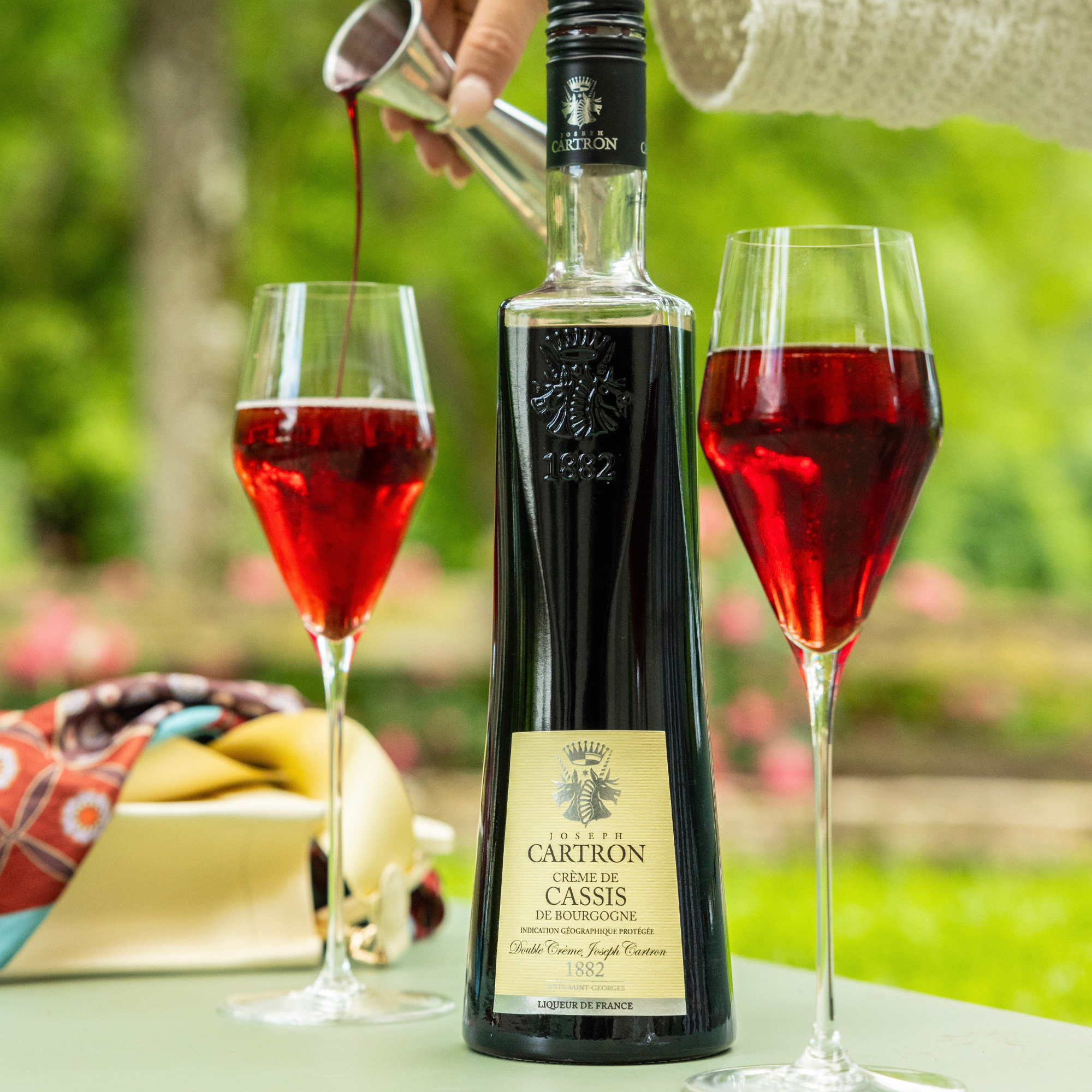Bottle of Cartron Creme de Cassis with two glasses being filled outdoors.