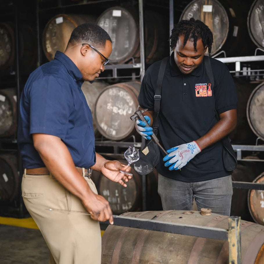 Two men working with a tool in front of barrels, likely in a brewery or distillery setting.