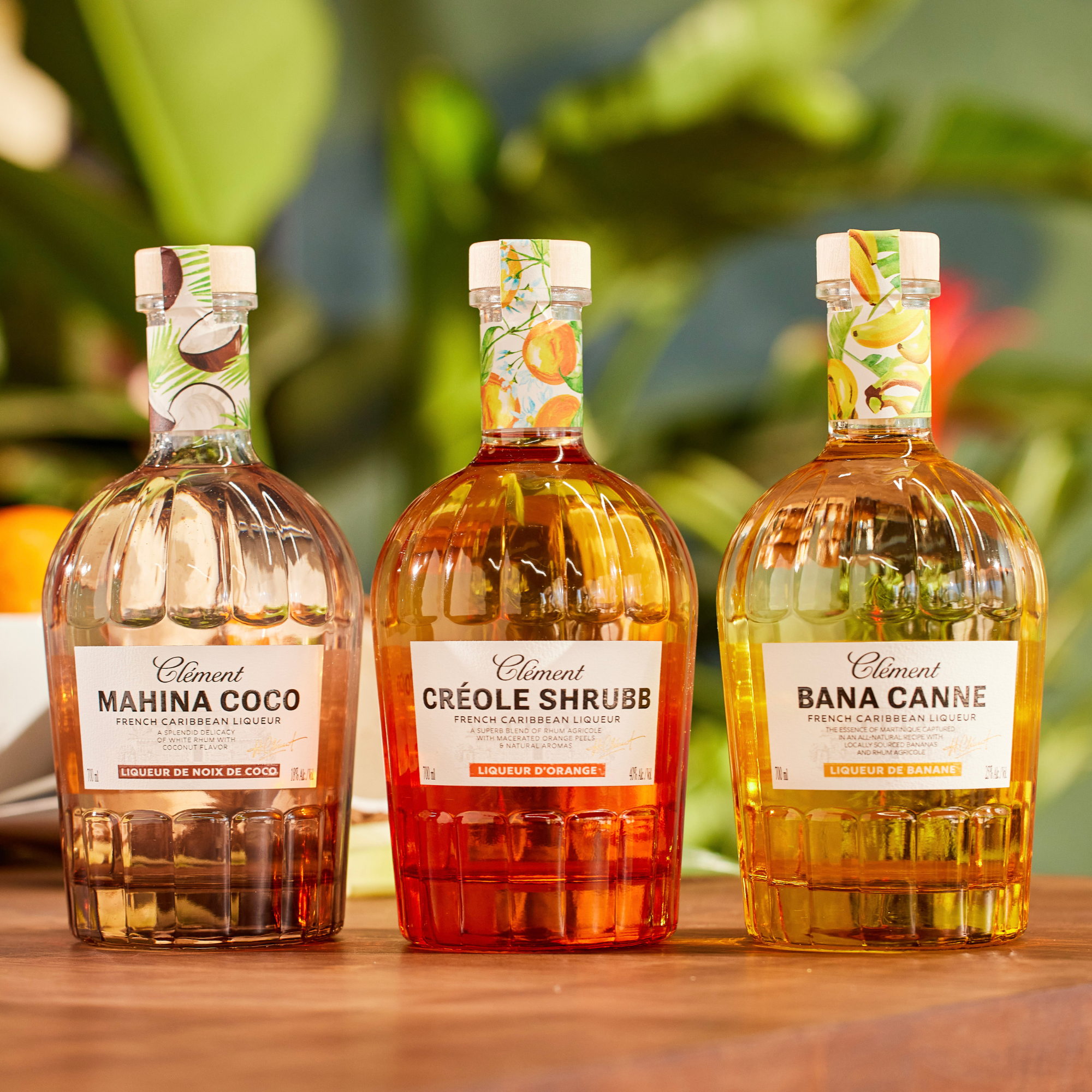Three bottles of Clément Mahina Coco, Créole Shrubb, and Bana Canne on a wooden surface with a blurred green background.