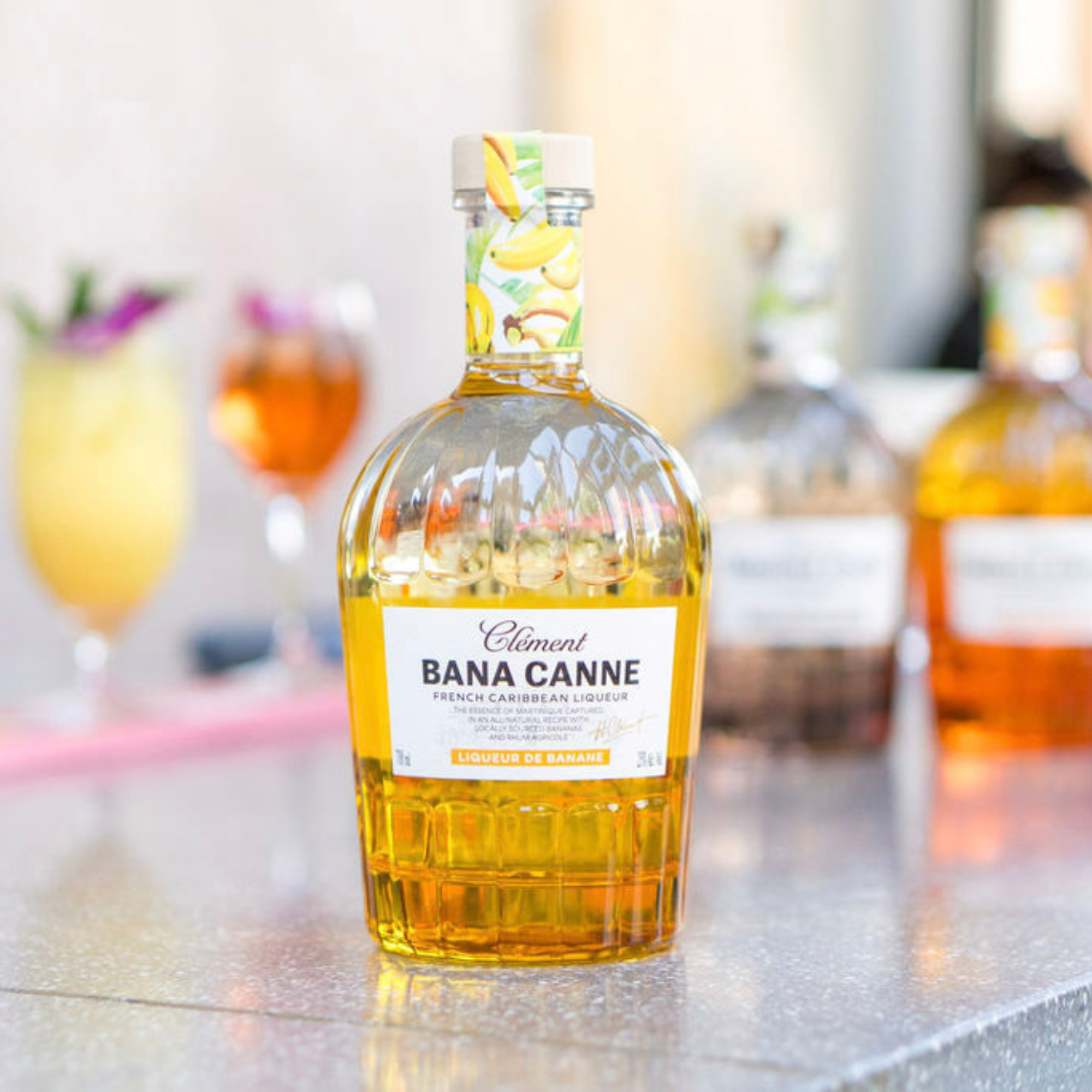 Bottle of Clément Bana Canne on a bar counter with blurred cocktails in the background