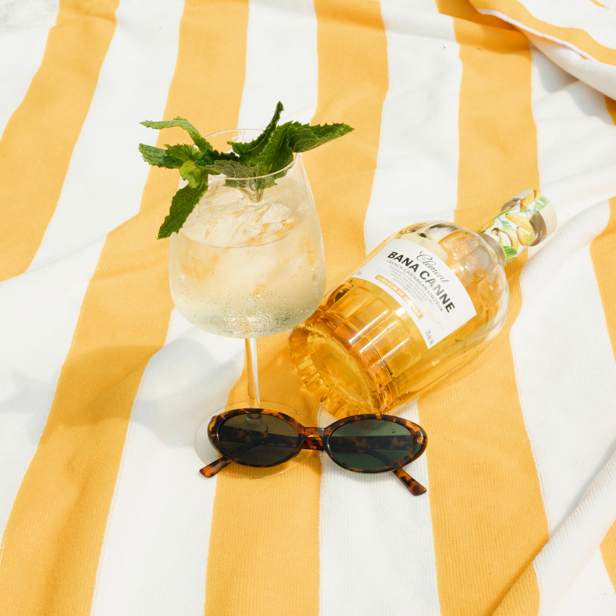 Bottle of Banca Cacique with a cocktail and sunglasses on a yellow and white striped fabric
