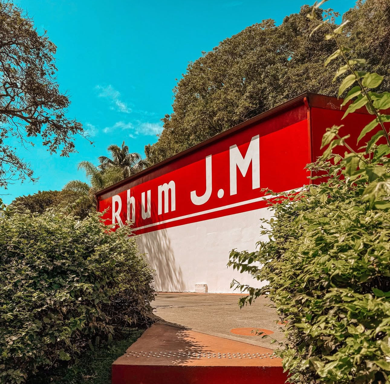 Red sign with 'Rhum J.M' on a building surrounded by trees and greenery.