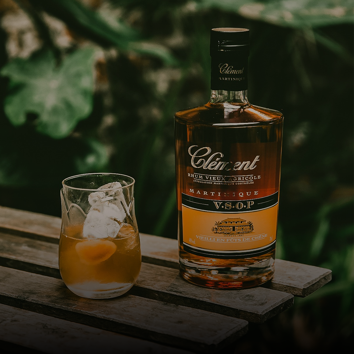 Bottle of Clement VSOP rum with a glass of cocktail on a wooden surface with a blurred green background