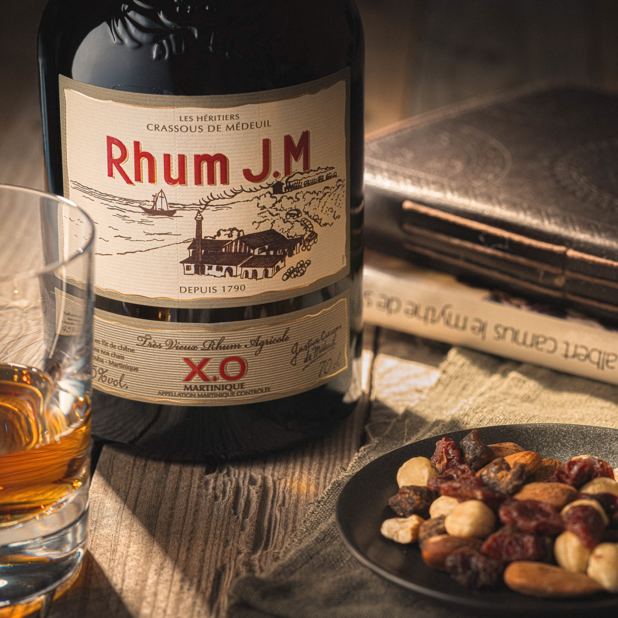 Rhum J.M XO rum bottle with a glass of rum and snacks on a wooden table.